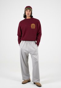 Burgundowy sweter z złotym emblematem, w parze z jasnoszarymi spodniami w stylu wide-leg. Modelka nosi zielono-czerwony czapkę w paski oraz brązowe buty.