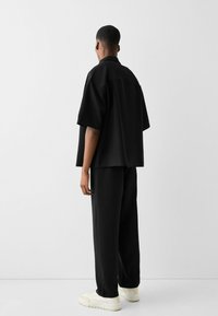 Übergroßes schwarzes kurzärmeliges Shirt mit geradem Schnitt, kombiniert mit lockeren schwarzen Hosen und weißen Schuhen, die ein minimalistisches Design betonen.