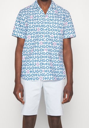 Homme portant une chemise à manches courtes boutonnée recouverte de texte « HUGO » bleu et violet avec un short blanc jusqu'aux genoux.