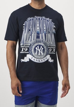 Mężczyzna noszący granatowy t-shirt z napisem „New York Yankees 1903” i grafiką o tematyce baseballowej w białych i szarych tonach.