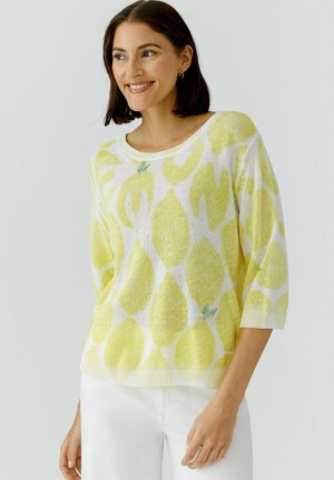 Femme souriante aux cheveux courts bruns portant un pull blanc orné de gros citrons jaunes et un pantalon blanc sur un fond uni.
