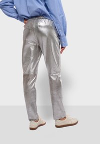 Glanzende zilveren metalen broek met een achterwaistband, twee zakken en rechte pijpen, gecombineerd met beige sneakers en een blauw gestreept shirt.