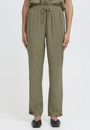 Persona che indossa pantaloni con coulisse in vita elasticizzata di colore verde oliva e ballerine nere con punta, in piedi contro uno sfondo chiaro e uniforme.