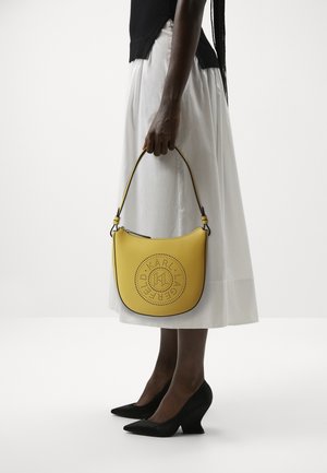 Sac à main en cuir jaune de forme croissant, avec un design de logo perforé et des finitions noires contrastantes. Porté avec une jupe blanche et des chaussures compensées noires.