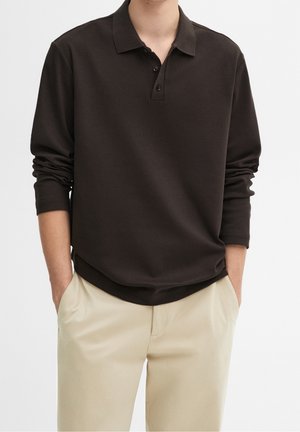 Person trägt ein dunkelbraunes langärmeliges Poloshirt mit drei Knöpfen und beigefarbene Hose, Hände in den Taschen, vor einfachem Hintergrund.