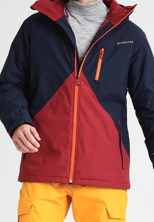 Homme portant une veste zippée bicolore bleu marine et rouge avec des détails de fermeture éclair orange et un pantalon cargo jaune moutarde.