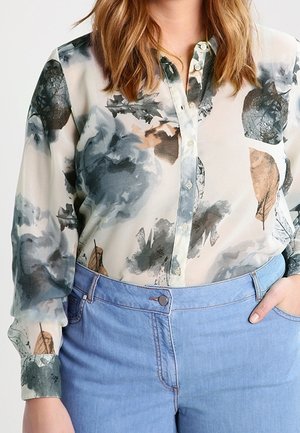 Femme portant un chemisier transparent à motif floral boutonné, rentré dans un jean taille haute bleu clair, main dans la poche.