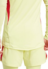 Limoengroene sporttop met rode zijaccenten en een mesh structuur; gecombineerd met bijpassende shorts. Bevat "AEROREADY" branding aan de zoom.