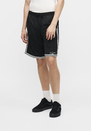 Άνδρας που φοράει μαύρο αθλητικό σορτς από διχτυωτό ύφασμα με λευκή διακόσμηση, λευκές κάλτσες, μαύρα αθλητικά παπούτσια και λευκό αμάνικο μπλουζάκι, στέκεται μπροστά σε λευκό φόντο.