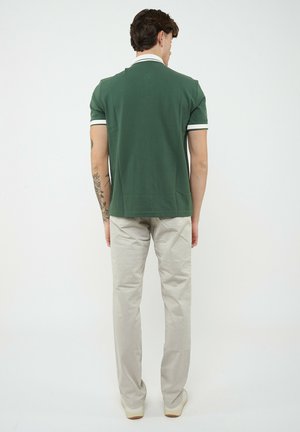 Uomo con polo verde e pantaloni beige, mostrato di spalle, in piedi contro uno sfondo bianco.