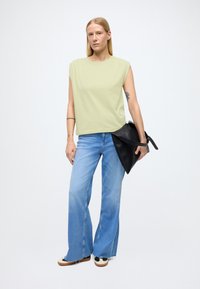 Haut vert clair sans manches associé à un jean évasé bleu. Le modèle tient un sac à main noir. Chaussures blanches et noires avec une semelle épaisse.