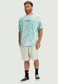 Light blue tie-dye t-shirt with short sleeves and a round neck, featuring black "Quiksilver" text. Worn with light shorts and black sneakers.