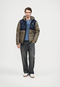 Padded jakna s khaki in temno modro barvno shemo, z zadrgo, kapuco in teksturirano tkanino z geometrijskim vzorcem.