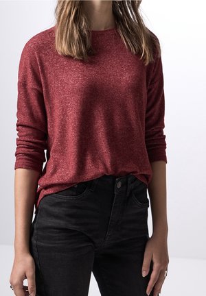 Femme portant un pull bordeaux à manches longues rentré dans un jean noir taille haute, debout devant un fond clair uni.