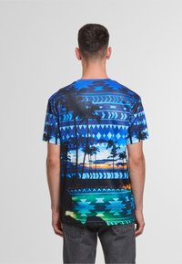 Short-sleeve blue graphic t-shirt featuring geometric patterns, palm trees, a sunset, and gradient colors from dark to light blue.