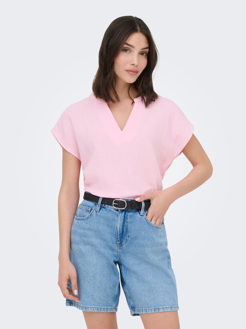 Femme aux cheveux mi-longs et noirs portant un haut rose clair à manches courtes et un short en jean taille haute, posant avec une main dans la poche.