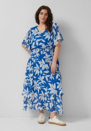 Kobieta stojąca przy szarej ścianie, ubrana w niebiesko-białą sukienkę w kwiaty i białe trampki, ręce swobodnie opuszczone wzdłuż ciała.