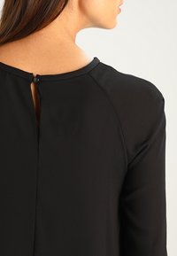 Blusa nera con scollo rotondo, realizzata in un tessuto testurizzato, con cuciture sulle spalle e chiusura a bottone sul retro per una vestibilità sicura.
