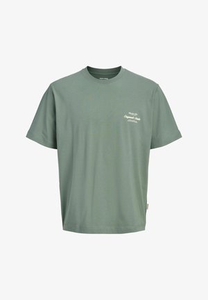 Garçon portant un sweat à capuche zippé bleu marine, un t-shirt vert, un pantalon marron et des chaussures beiges, debout avec une main dans la poche, regardant sur le côté.