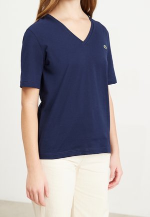 Persona que lleva una camiseta azul marino de manga corta con cuello en V y un pequeño logo de cocodrilo verde en el pecho, junto con pantalones color crema.