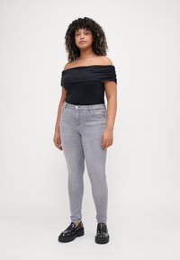 Fekete, váll nélküli felső, drapírozott anyagból, világosszürke skinny farmer és fekete platform cipők díszes szegecsekkel. Semleges háttér.
