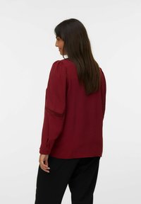 Bordeaux blouse met geplooide pofmouwen en kantdetails aan de manchetten, gemaakt van gestructureerde stof; gedragen met zwarte broek.