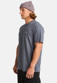 Grå kortærmet t-shirt med rund hals, der har et subtilt logo på brystet og en glat bomuldsstof. Sættes sammen med en let strikket bøllehat.