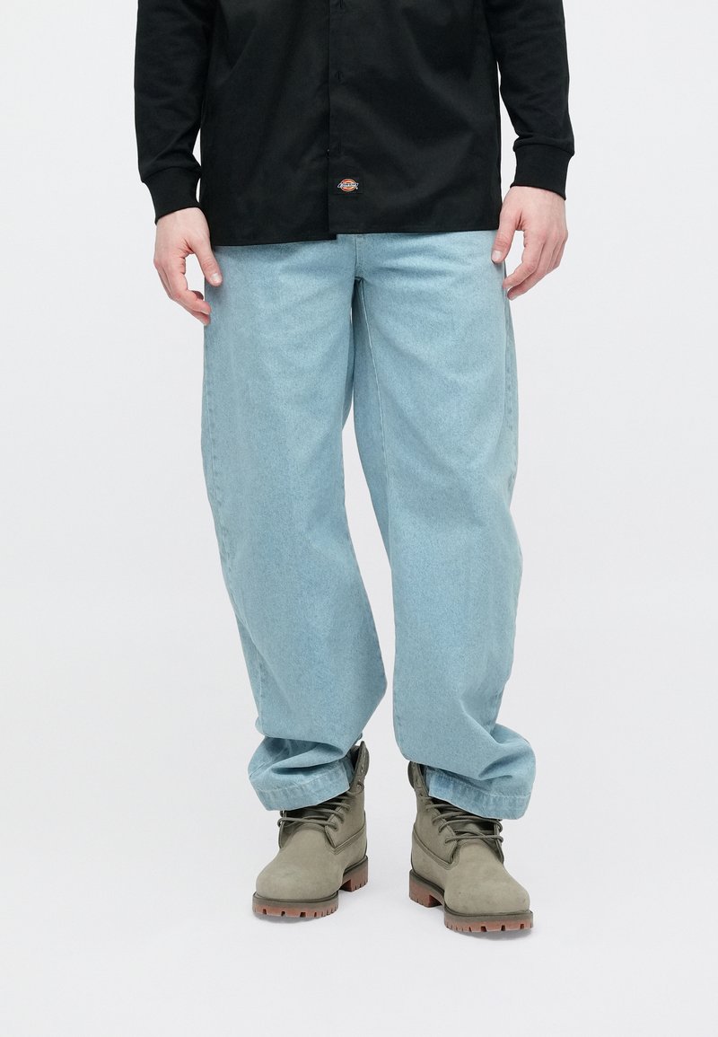 Jean large couleur bleu clair avec une texture délavée, associé à des bottes vertes. La chemise est noire, à manches longues, avec un petit logo.