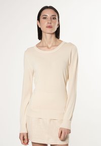 Maglione beige a maniche lunghe con design aderente, scollo rotondo, e polsini e orlo a costine. Texture liscia, dettaglio hardware minimale.