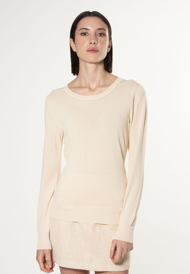 Maglione beige a maniche lunghe con design aderente, scollo rotondo, e polsini e orlo a costine. Texture liscia, dettaglio hardware minimale.