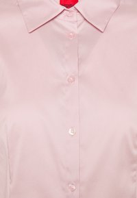 Blusa rosa chiaro con colletto classico, composta da cinque bottoni e tessuto dalla texture liscia e lucida. Etichetta del marchio visibile sul retro del colletto.