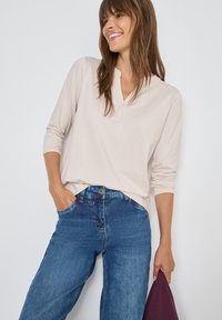 Top lungo beige chiaro a maniche lunghe con scollo a V e una vestibilità rilassata, abbinato a jeans dritti blu medio. Il modello tiene un oggetto di un borgogna scuro.