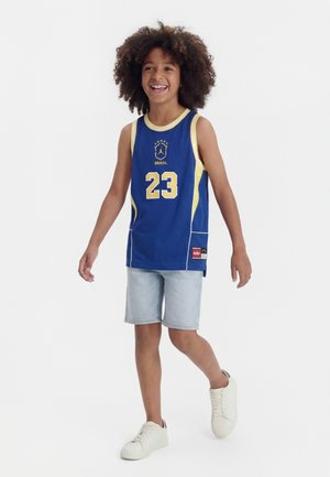 Niño con cabello rizado sonriendo, caminando, vestido con camiseta de baloncesto de Brasil azul y amarilla número 23, pantalones cortos de mezclilla clara y zapatillas blancas.