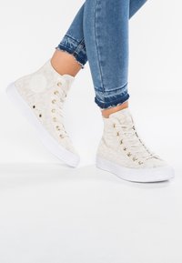 Wysokie sneakersy wykonane z teksturowanego beżowego materiału z złotymi oczkami i białą gumową podeszwą. Posiadają okrągłe logo po boku.