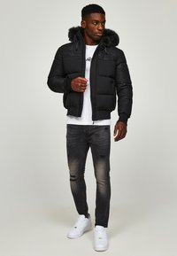Chaqueta de abrigo negra con capucha forrada de piel, cierre de cremallera y puños acanalados, combinada con jeans grises desgastados y zapatillas blancas.