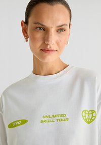 Witte katoenen t-shirt met groene grafische tekst "UNLIMITED SKULL TOUR" en een hartontwerp met een schedelicoon, ronde halslijn, korte mouwen.