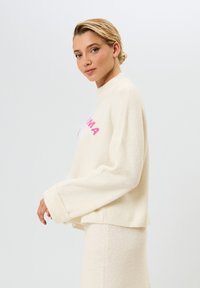 Witte cropped sweater met roze tekst, wijde mouwen en een zachte textuur, gecombineerd met bijpassende lichte stof. Eenvoudig, modern ontwerp.