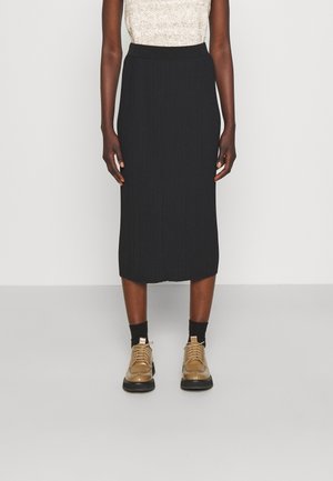 Zwarte geribbelde midi-rok met een elastische tailleband, gecombineerd met tan geknoopte schoenen en zwarte sokken, weergegeven tegen een neutrale achtergrond.