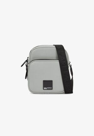 Pelēka mākslīgās ādas sajūga soma ar taisnstūra formu, ar melnu regulējamu siksnu un kontrastējošu logotipa plāksni priekšpusē.