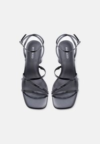 Μαύρα σανδάλια με λουράκια, τετράγωνες μύτες και αγκράφες στον αστράγαλο, κομψό δερμάτινο φινίρισμα, επίπεδη σόλα, λογότυπο της μάρκας στις εσωτερικές σόλες, σε λευκό φόντο.