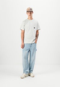 Gray short-sleeve t-shirt with a chest pocket, light blue baggy jeans, and beige sneakers, accessorized with a tan cap featuring a logo.