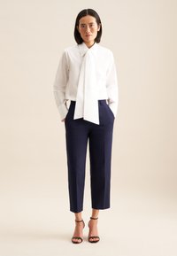 Camicia bianca a maniche lunghe con cravatta, abbinata a pantaloni blu navy alla caviglia e tacchi neri con cinturini. Tessuto liscio con una vestibilità su misura.