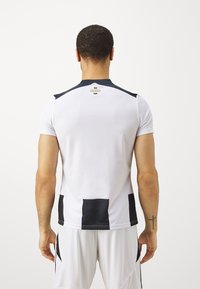 Maglietta sportiva bianca con accenti neri, maniche corte e scollatura rotonda. Presenta un logo sulla schiena e motivi rettangolari neri sui lati.