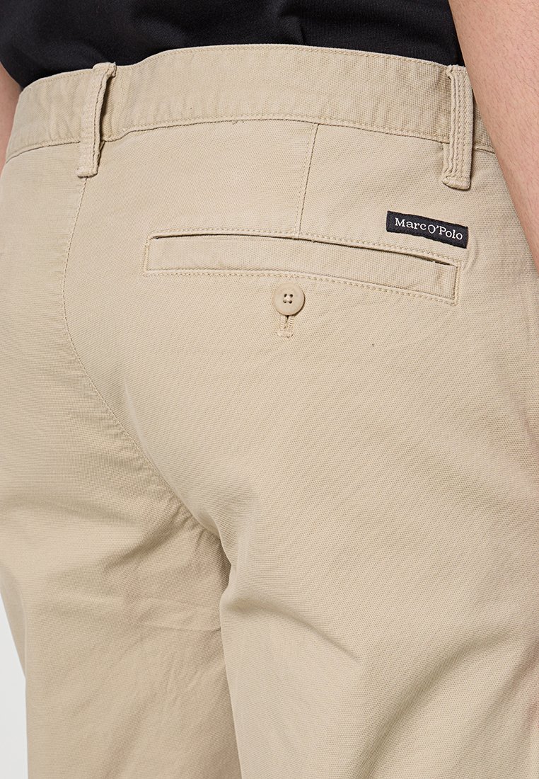 Beżowe bawełniane spodnie z tylną kieszenią zapinaną na guzik i widoczną metką marki, o gładkiej fakturze i dopasowanym kroju.