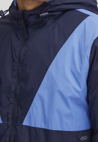 Veste en nylon bleu marine et bleu clair avec une fermeture éclair, présentant un bloc de couleurs diagonal, une capuche à cordon de serrage et un petit patch logo.