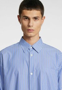 Svītraina krekla ar gaiši zili baltām vertikālām svītrām, smailu apkakli, pogu aizdari priekšpusē un kreisā pusē krūšu kabatu.