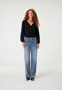 Schwarze Bluse mit Knopfverschluss und langen Ärmeln, kombiniert mit hellblauen weit geschnittenen Jeans und schwarzen Sandalen mit weißen Akzenten. Rote Perlenkette inklusive.