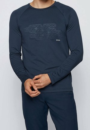 Mężczyzna noszący granatową koszulę z długim rękawem, na której powtarza się wytłaczany napis "BOSS" oraz małe białe logo "BOSS" na piersi.