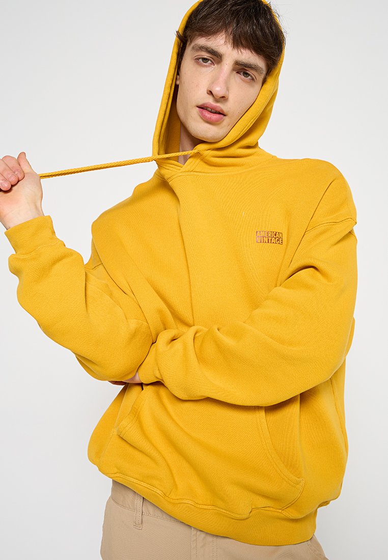 Geel hoodie met een trekkoordcapuchon, geribbelde boorden en een kangoeroezak. Bevat een klein "American Vintage" logo aan de voorkant. Zachte textuur.