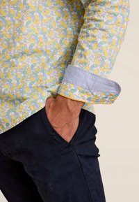 Langarmshirt met een bloemenpatroon van gele citroenen en groene bladeren op een lichtblauwe achtergrond. Blauwe gestructureerde manchetten. Donkere broek.